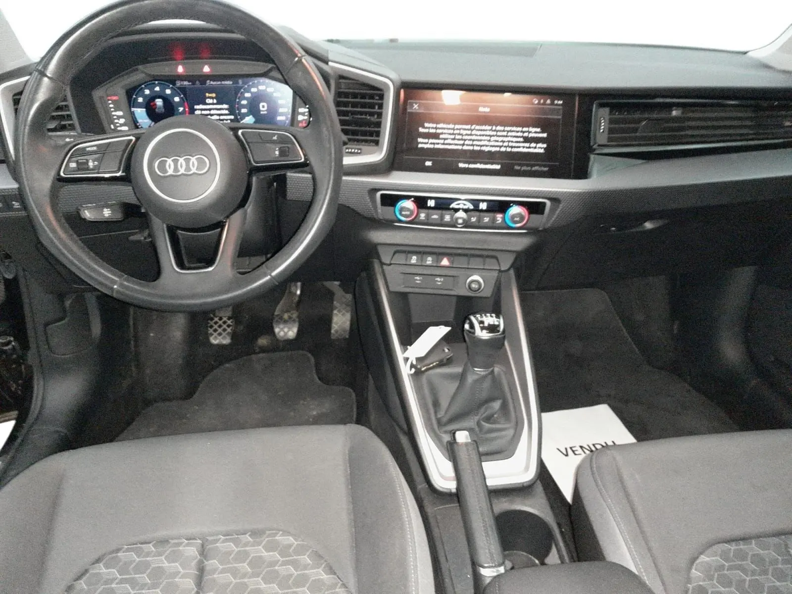 Vue intérieure avant de l'Audi A1 Sportback 2023 noire, avec volant multifonction, tableau de bord digital et levier de vitesse manuel.