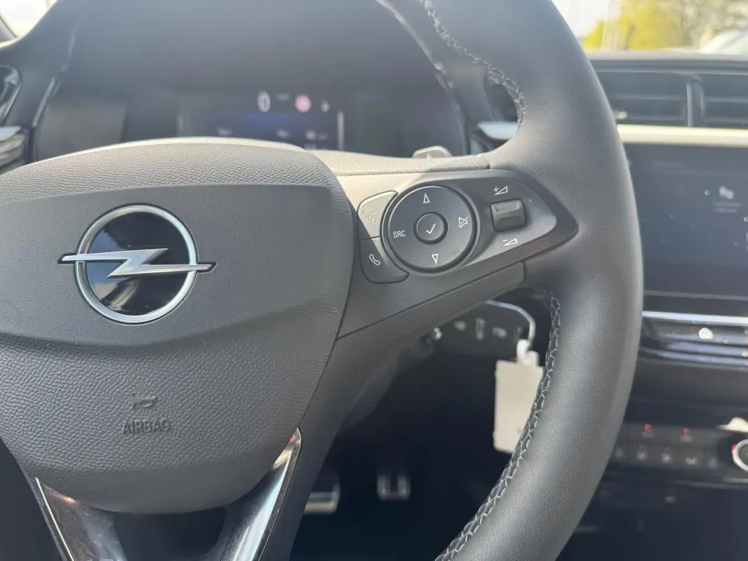Vue rapprochée du volant multifonctions noir de l’Opel Corsa 2025 avec commandes audio et tableau de bord flou.