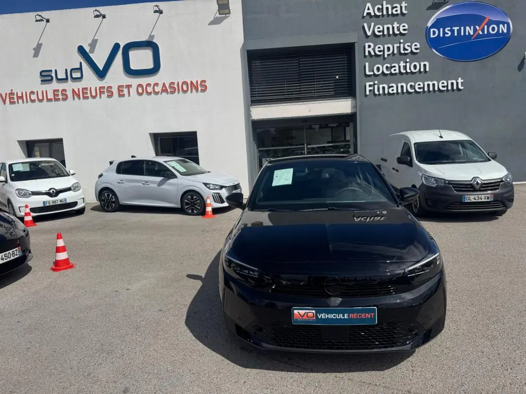 Opel Corsa noire vue de face, garée devant un concessionnaire avec panneaux de vente en arrière-plan.