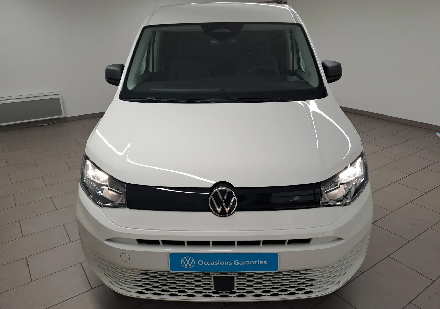 Vue avant d’un Volkswagen Caddy Cargo Maxi blanc candy 2026 avec calandre noire et phares allumés en intérieur showroom.