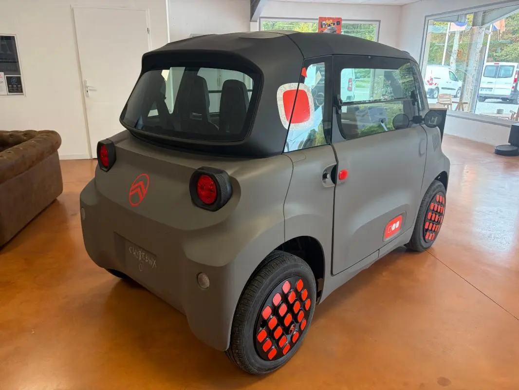 Vue 3/4 arrière droite de la Citroën Ami Spicy 2025 gris mat avec détails rouges sur roues et carrosserie.