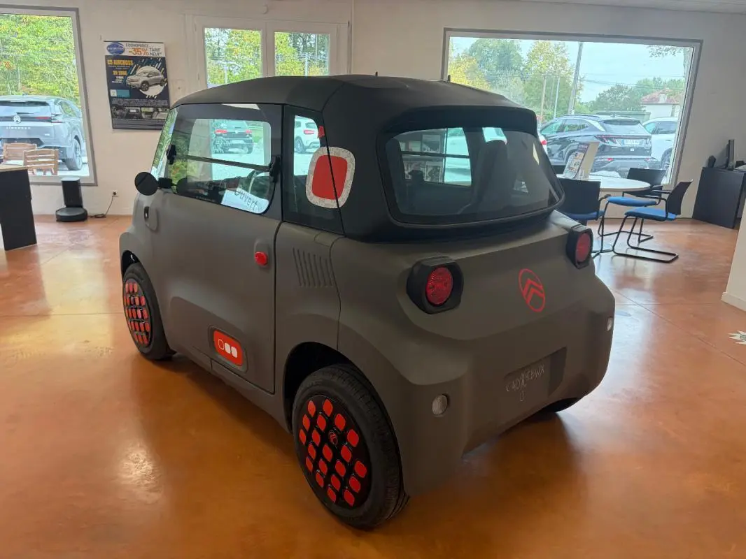 Vue 3/4 arrière droite de la Citroën AMI Spicy 2025 gris avec détails rouges distinctifs et toit noir en intérieur showroom.