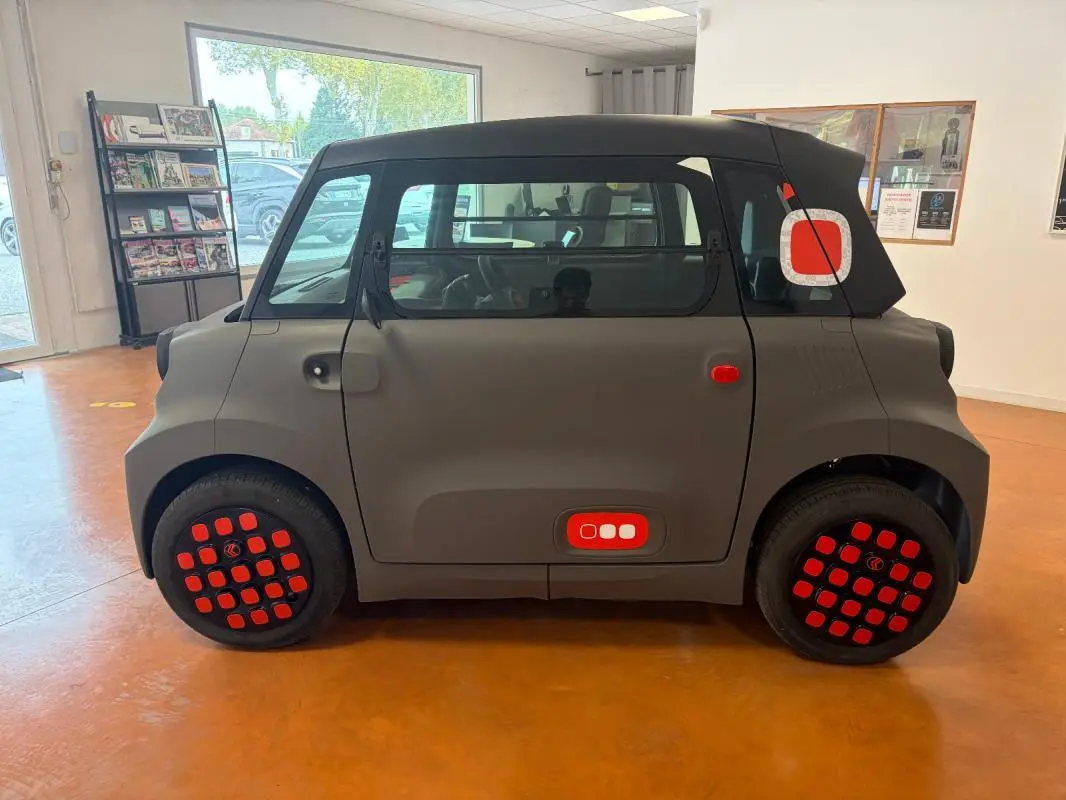 Vue de profil côté gauche d'une Citroën Ami Spicy 2025 gris mat avec détails rouges sur les roues et la porte arrière.