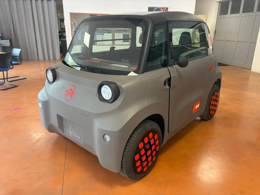 Vue 3/4 avant droite du Citroën Ami Spicy gris avec détails rouges sur jantes et logo, dans un intérieur lumineux.