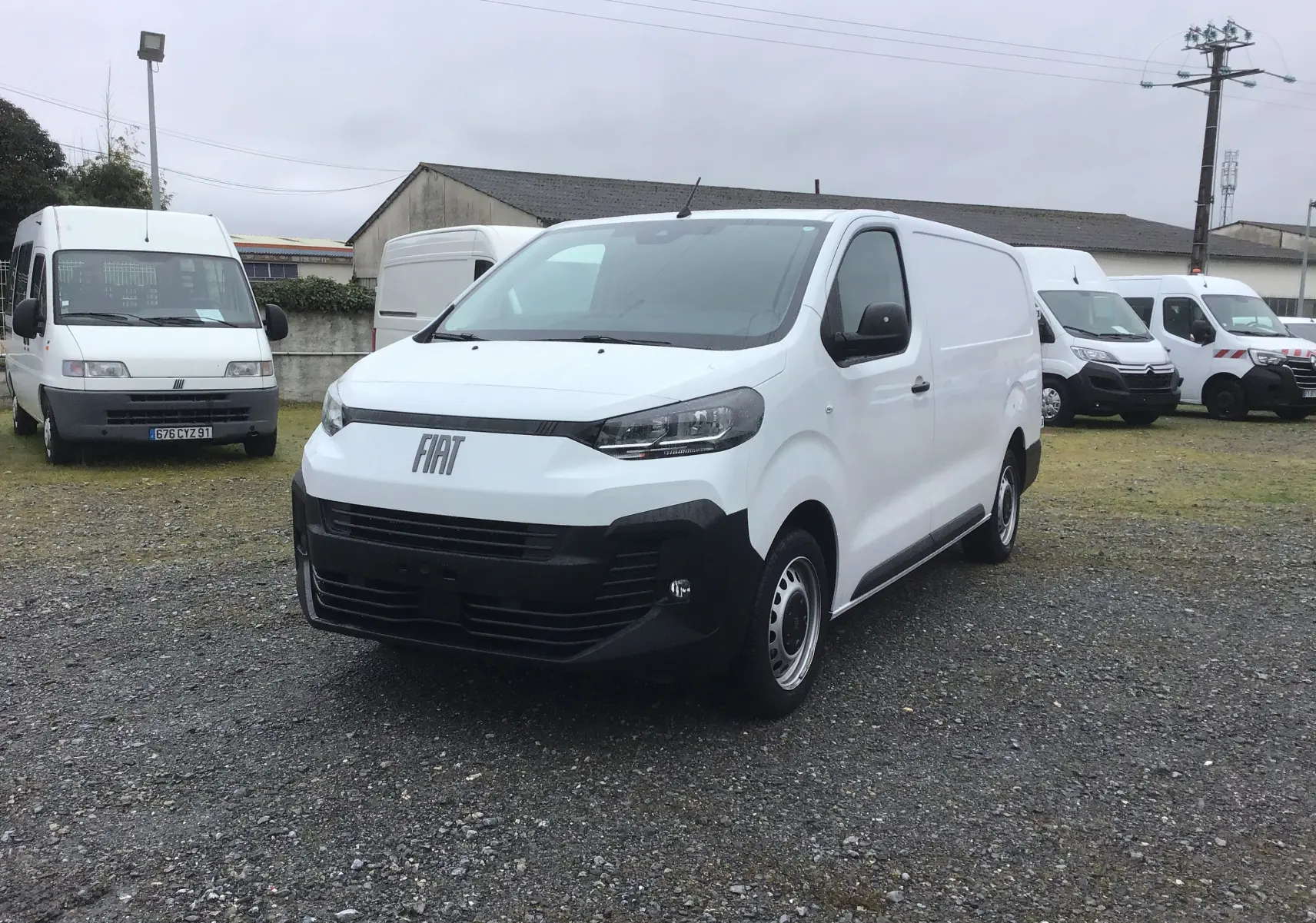 Vue 3/4 avant d’un FIAT Scudo Fourgon blanc 2024 avec pare-chocs noir et jantes tôles, stationné sur un sol gravillonné.