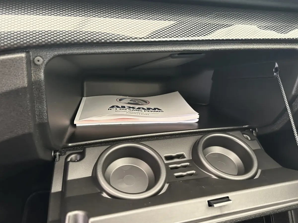 Gros plan sur la boîte à gants ouverte de l'AIXAM AMBITION blanche, avec porte-gobelets intégrés et manuel visible.