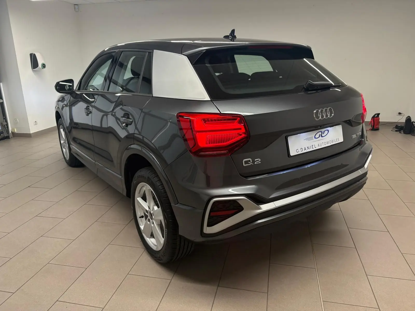 Audi Q2 35 TFSI S Line gris Daytona vu en 3/4 arrière droit, avec toit contrasté argent et feux arrière allumés.
