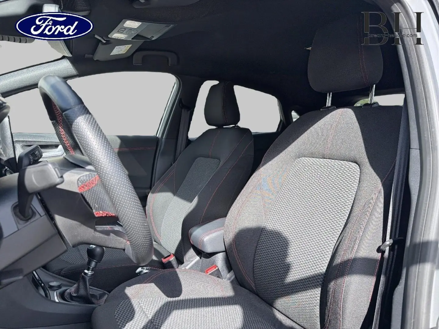 Vue intérieure côté conducteur du Ford Puma 2024, sièges tissu noir avec surpiqûres rouges et volant cuir perforé.