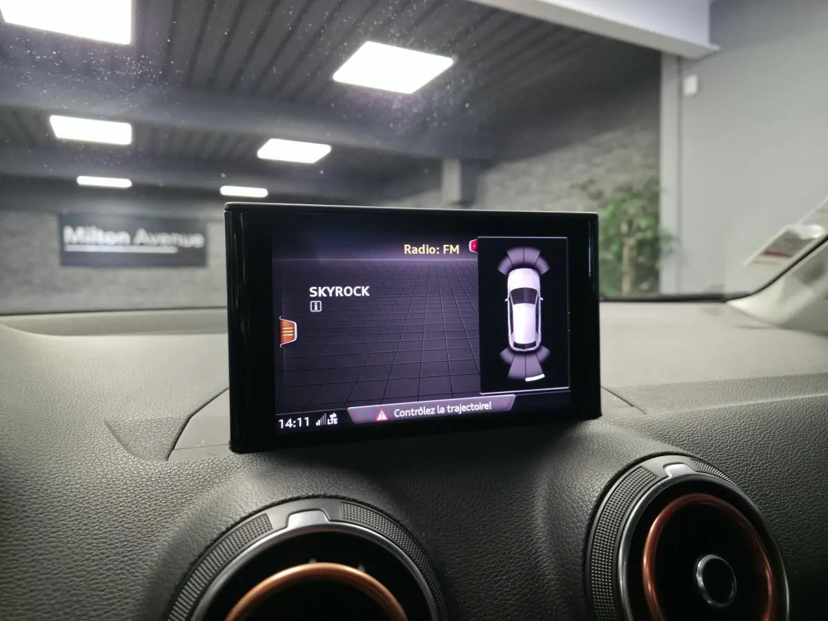 Écran MMI central affichant la radio FM et l'aide au stationnement dans l'Audi Q2 noire, vue intérieure avant.