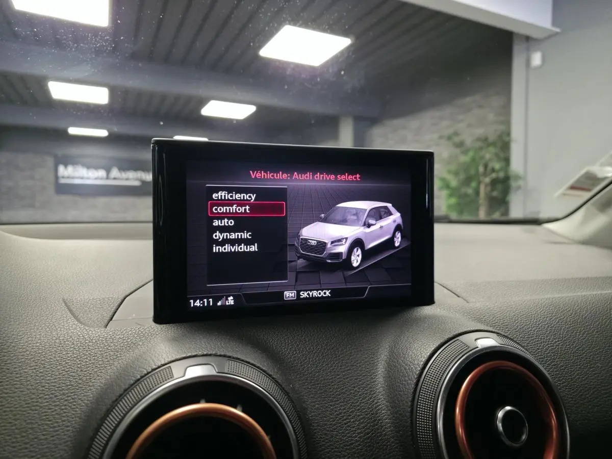 Écran MMI affichant Audi Q2 blanc en vue 3/4 avant, avec tableau de bord noir et aérateurs cerclés de rouge.