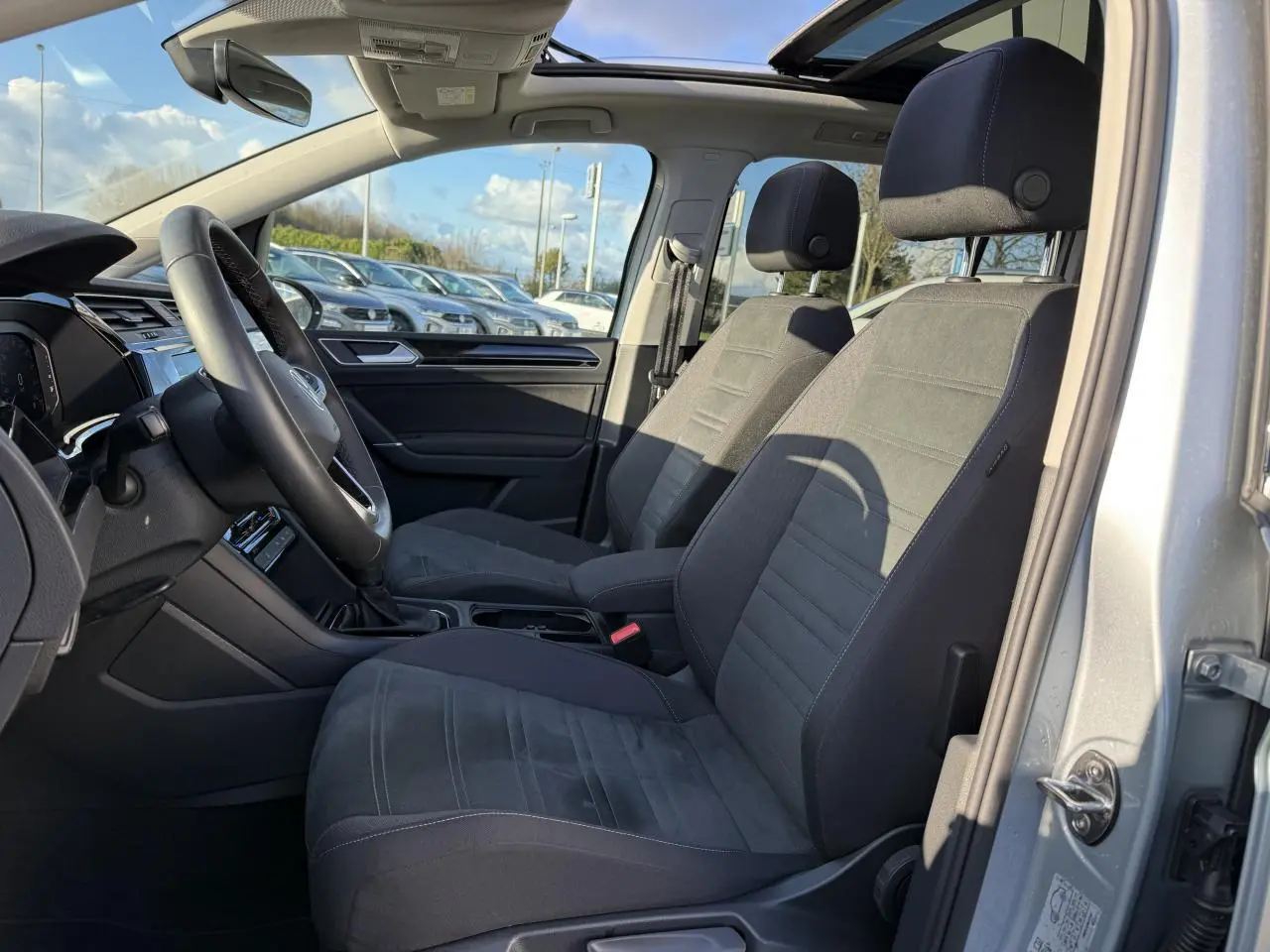 Vue intérieure côté conducteur du Volkswagen Touran argent Dolomite 2025 avec toit ouvrant panoramique et sellerie microfleece noire.