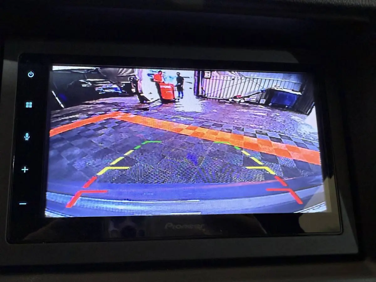 Vue écran caméra de recul côté conducteur d'une Aixam Ambition 0.5 Crossover Premium gris foncé, affichant le sol pavé et une barrière.