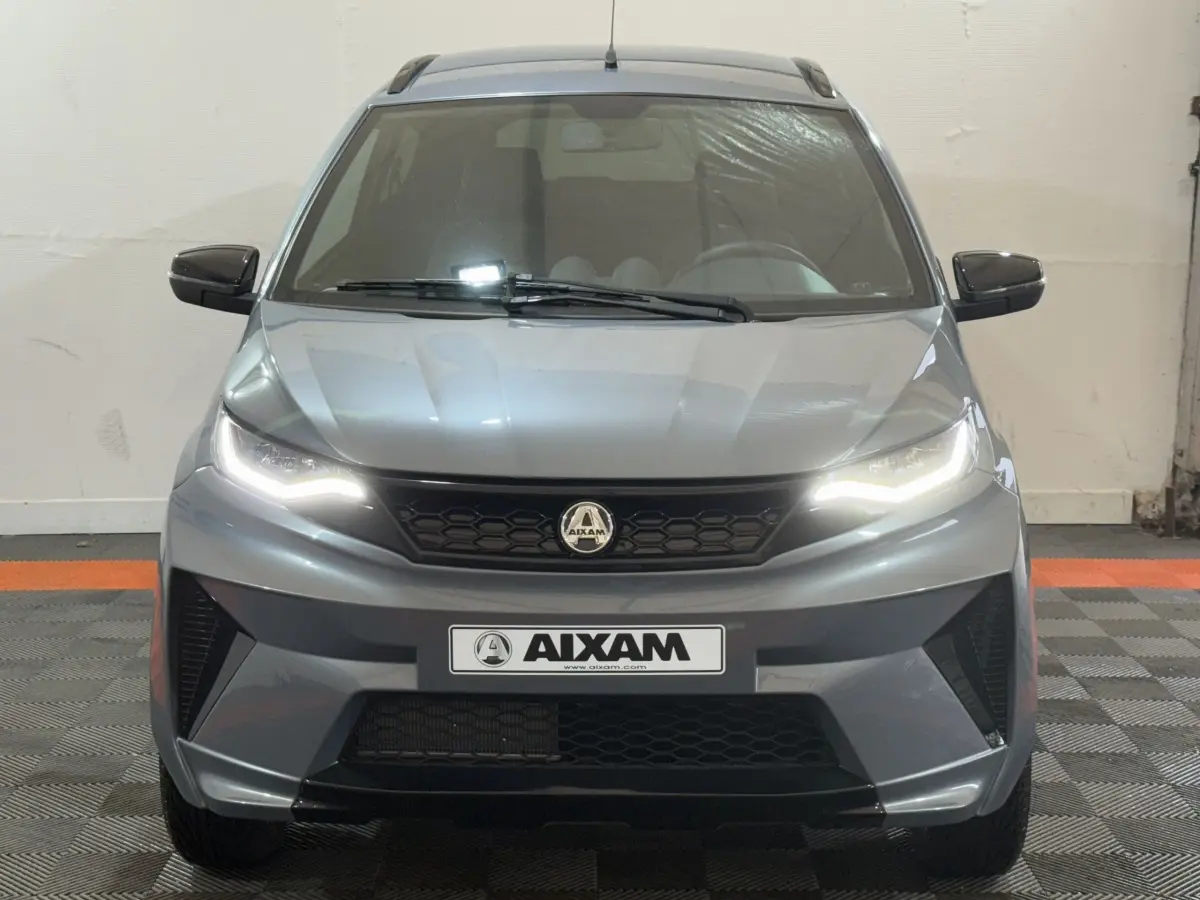 Vue frontale d'un Aixam Ambition 0.5 Crossover Premium gris foncé avec phares allumés et rétroviseur caméra côté gauche.