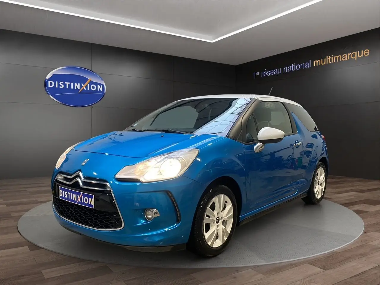 Citroën DS3 bleu avec toit blanc en 3/4 avant droit, phare allumé et jantes blanches visibles en showroom.