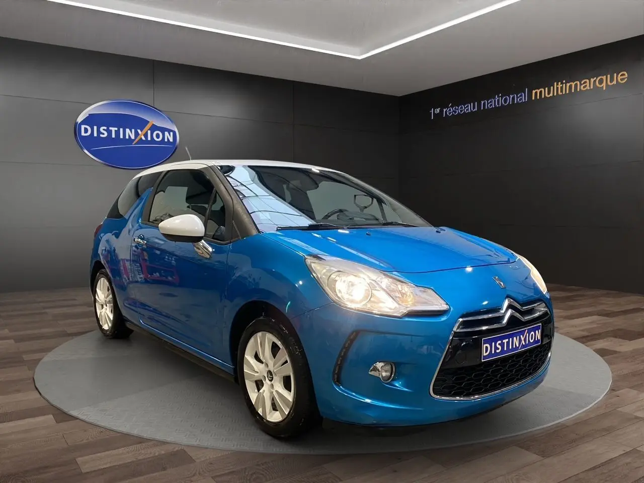 Citroën DS3 bleu avec toit blanc en 3/4 avant droit, phare allumé et jantes blanches visibles en intérieur showroom.