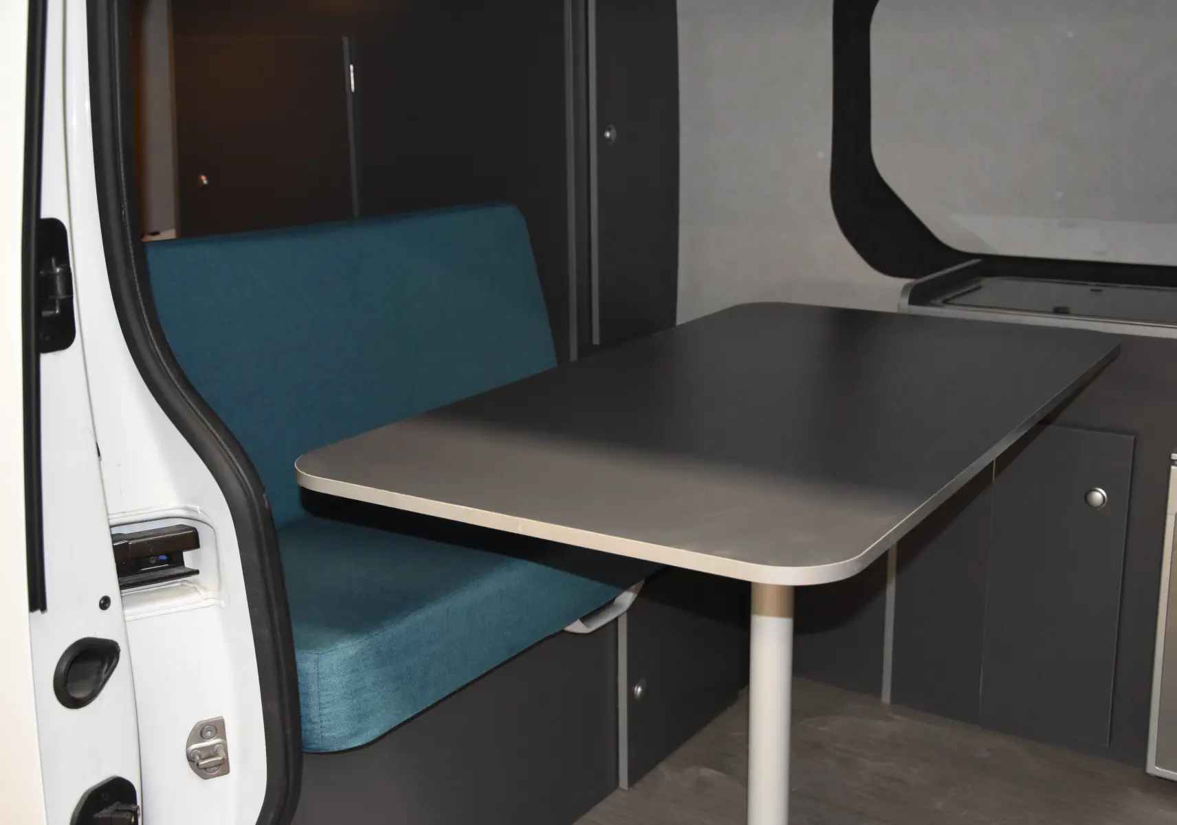 Coin repas intérieur avec banquette bleue et table grise dans un Renault Trafic Combi blanc, porte latérale ouverte.