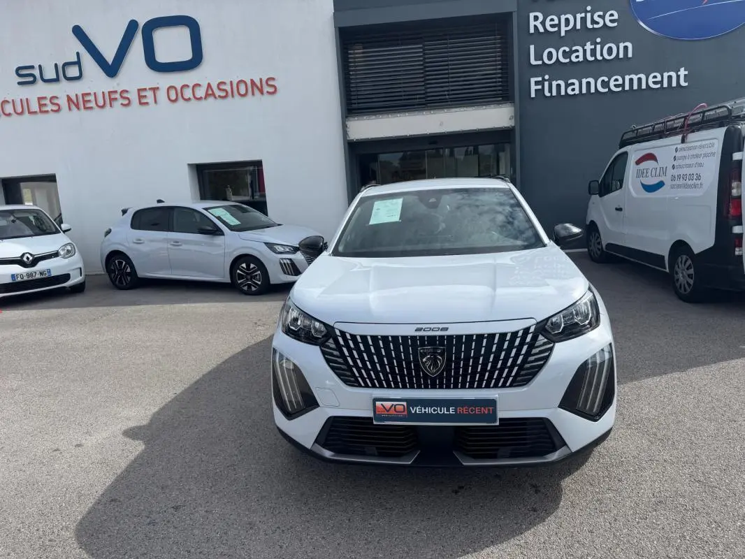 Vue avant d’un Peugeot 2008 blanc 2024 avec calandre noire et barres de toit, stationné devant un garage.