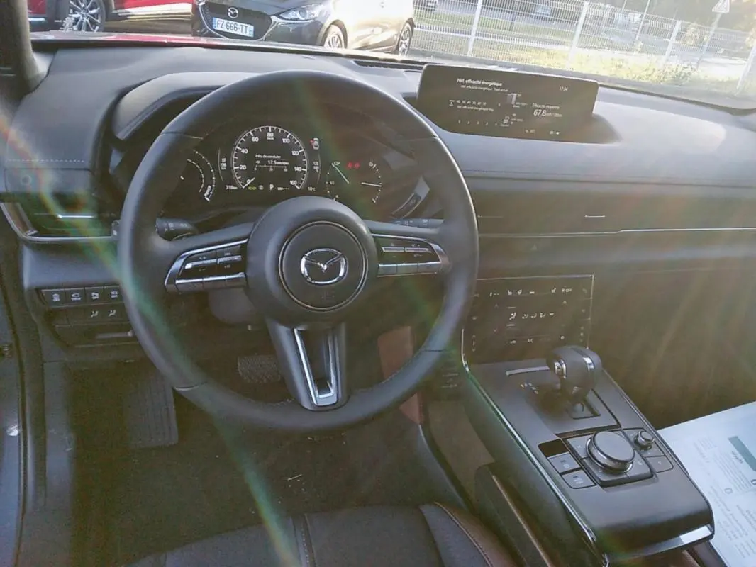 Vue intérieure du tableau de bord et volant du Mazda MX-30 gris, avec écran central et levier de boîte automatique visible.