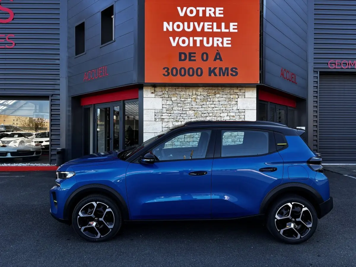 Profil côté gauche d'une Citroën C3 100 BVM PLUS bleu 2025 avec toit noir et jantes modernes, devant un bâtiment commercial.