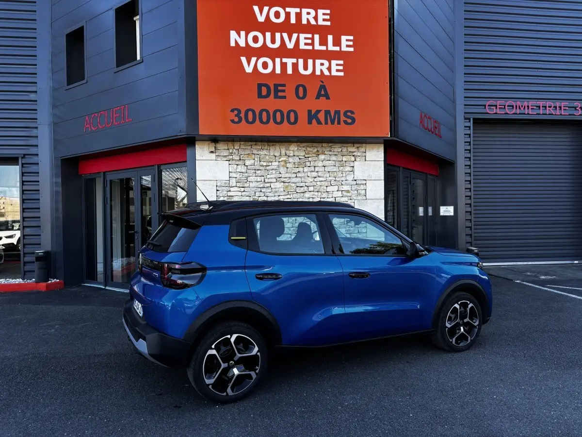 Vue de profil droit d'une Citroën C3 100 BVM PLUS bleu avec toit noir, roues alliage et feux arrière LED, devant un garage.