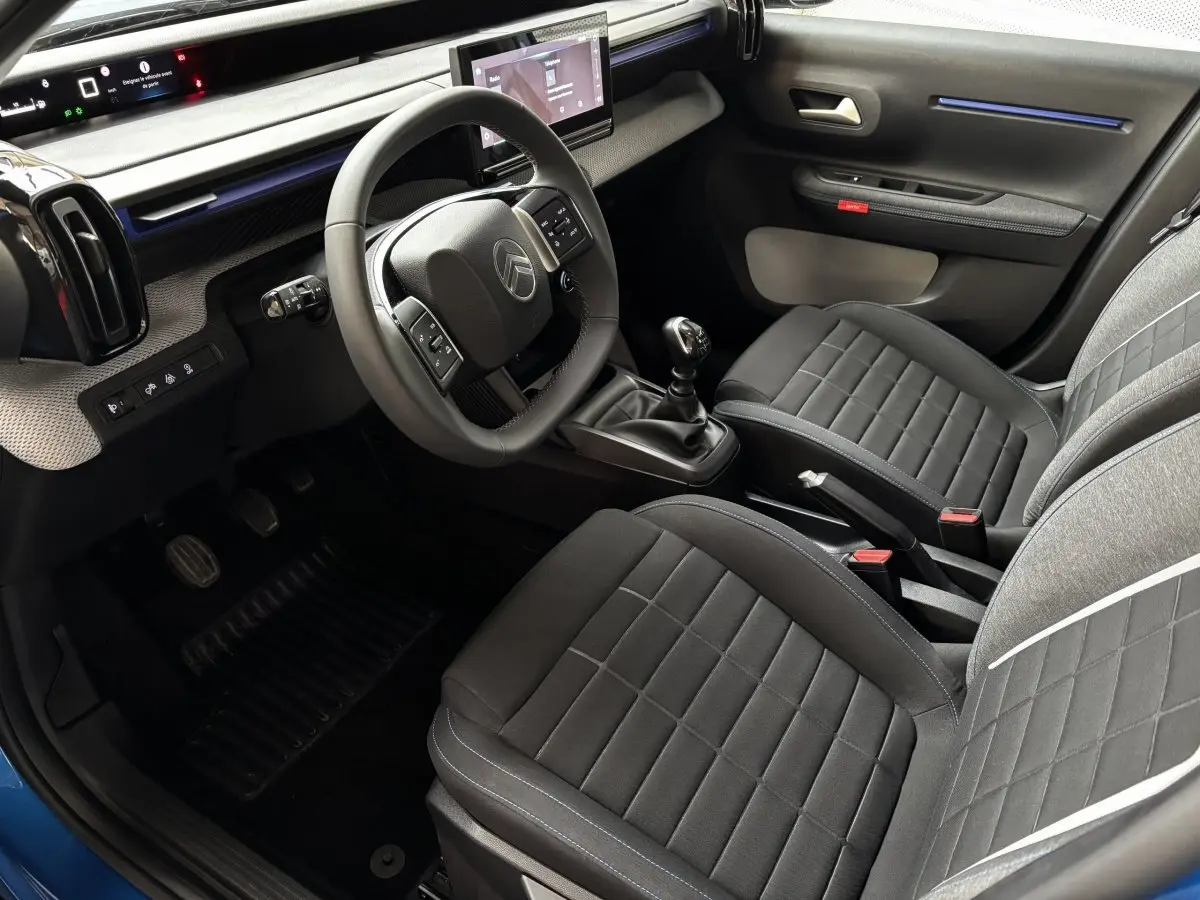 Vue intérieure côté conducteur de la Citroën C3 100 BVM PLUS 2025 avec sièges noirs et volant multifonctions.