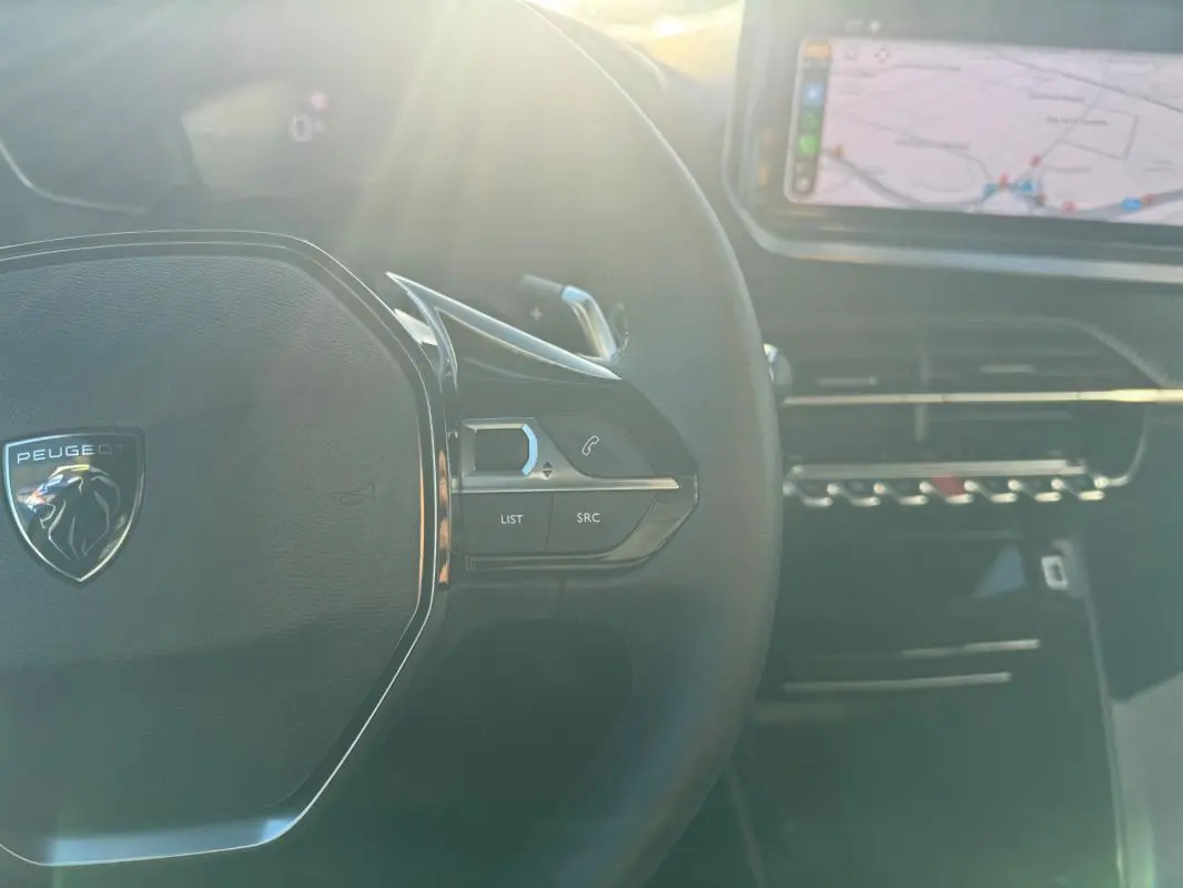 Vue rapprochée du volant noir du Peugeot 2008 Hybrid 136 gris avec commandes et écran tactile en arrière-plan.