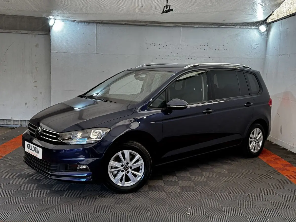 Volkswagen Touran bleu 3/4 avant droit en intérieur, avec jantes alliage et rails de toit chromés visibles.
