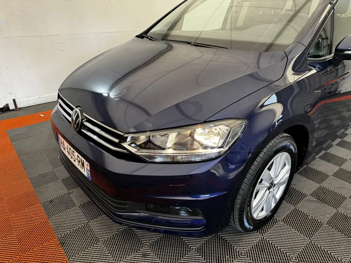 Vue 3/4 avant droit d’un Volkswagen Touran bleu 2024 avec phares allumés et jantes argentées.
