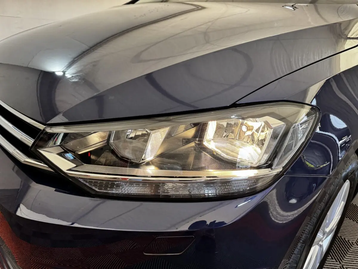Gros plan sur le phare avant droit d’un Volkswagen Touran bleu 2024, mettant en valeur la calandre chromée et la carrosserie brillante.