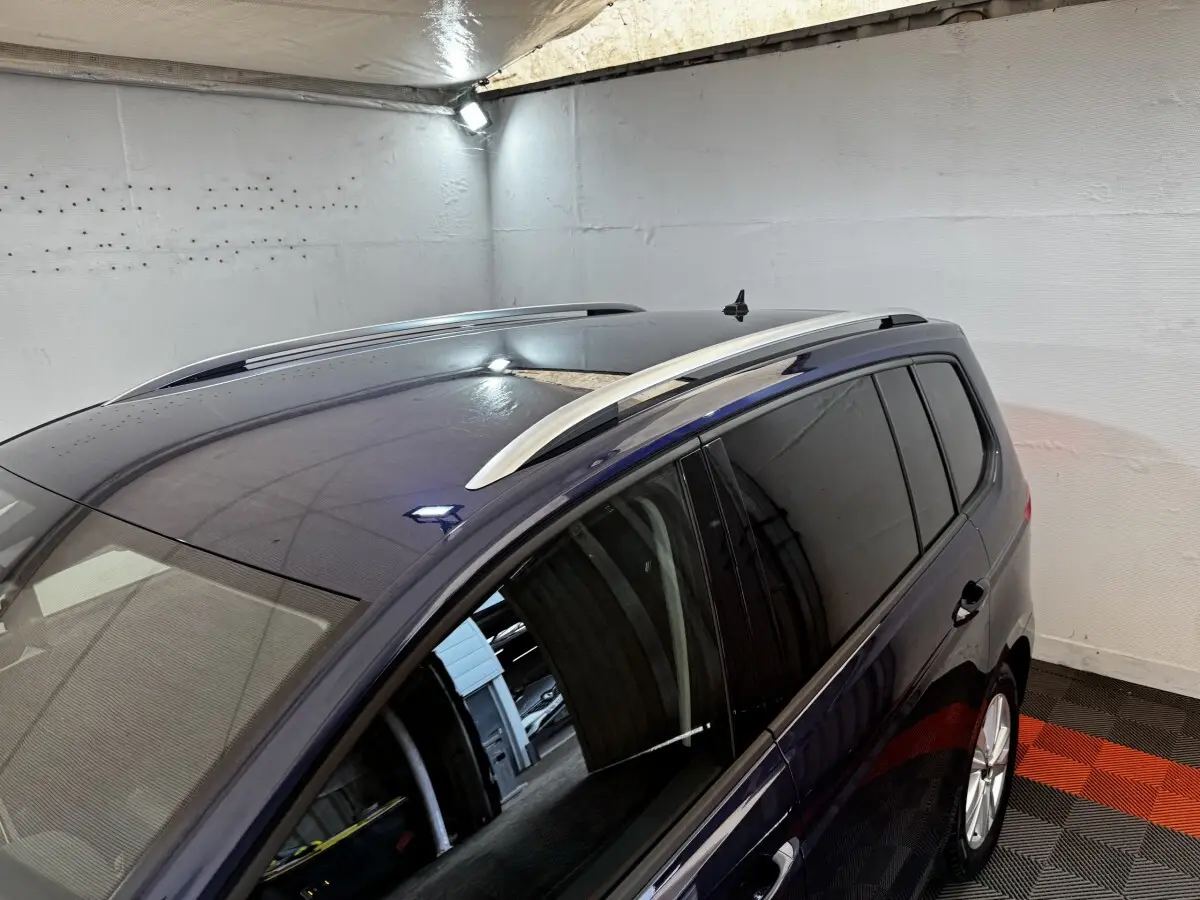 Vue latérale droite du Volkswagen Touran bleu 2024 avec barres de toit argentées et vitres teintées dans un garage.
