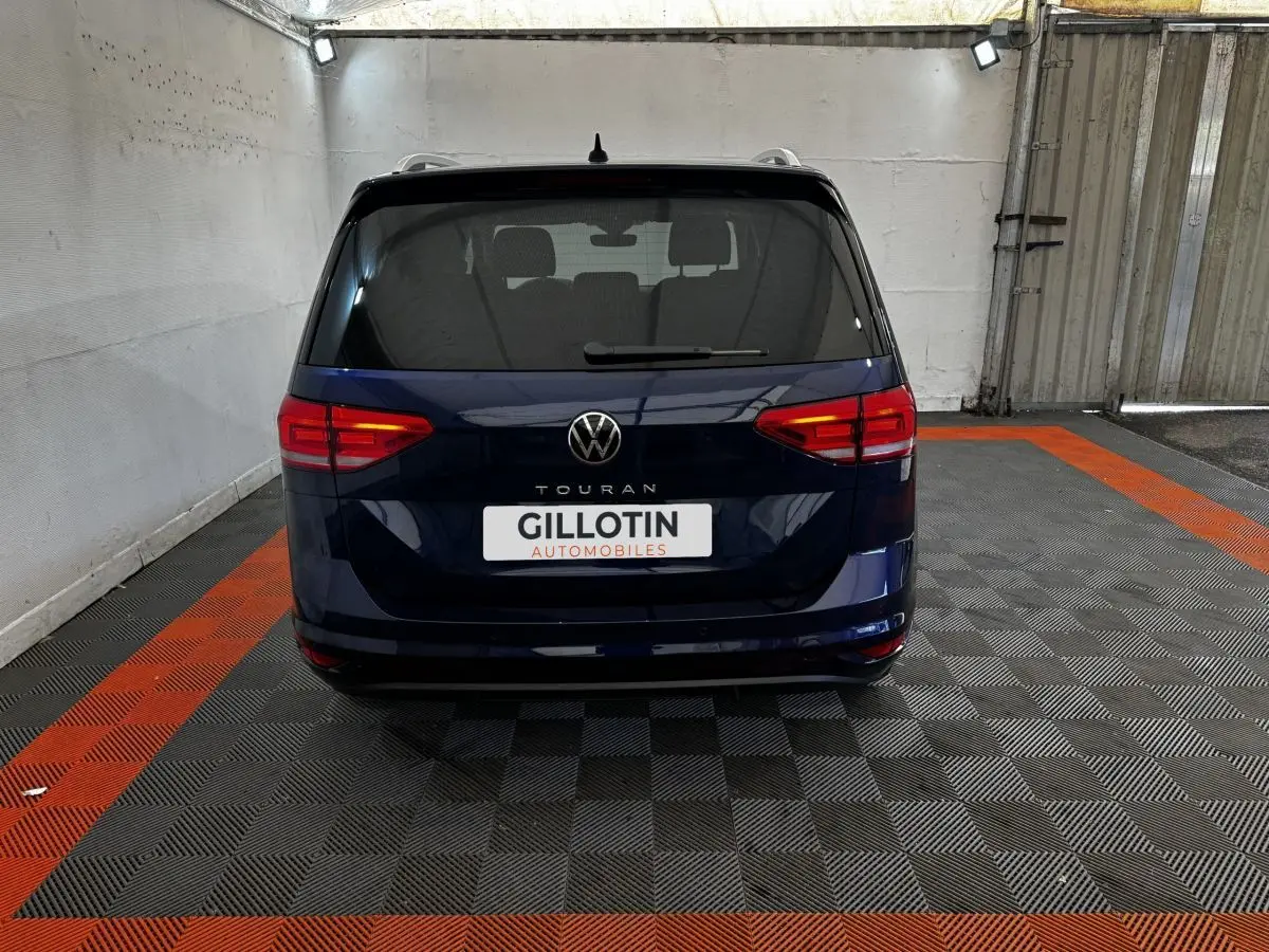 Vue arrière d'un Volkswagen Touran bleu 2024 garé sur un sol à damiers noir et orange dans un garage.