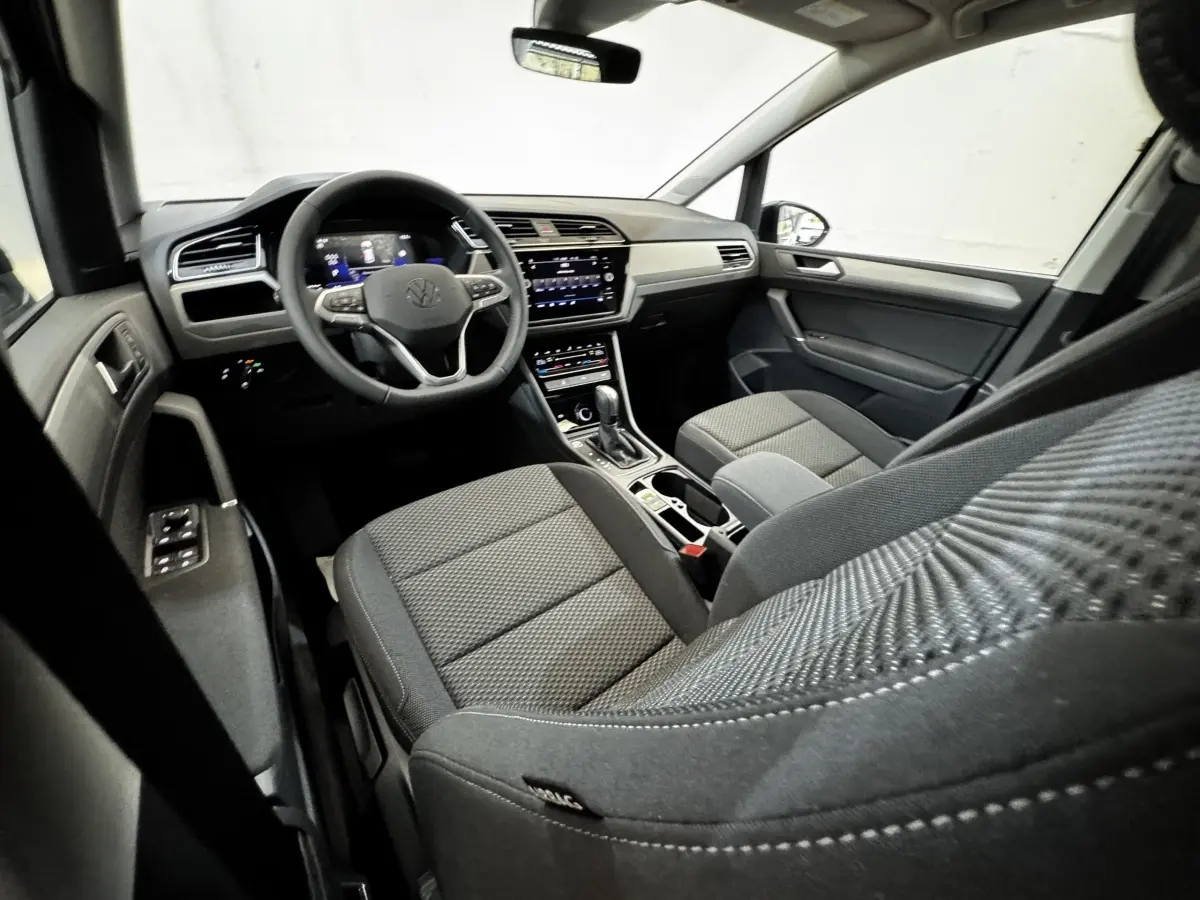 Vue intérieure côté conducteur du Volkswagen Touran 2024, montrant le tableau de bord et les sièges en tissu gris.