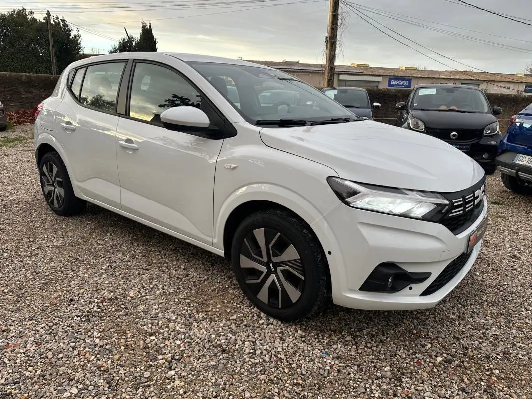 Vue 3/4 avant droit d'une Dacia Sandero TCe 90 Expression blanche avec jantes noires et phares allumés.