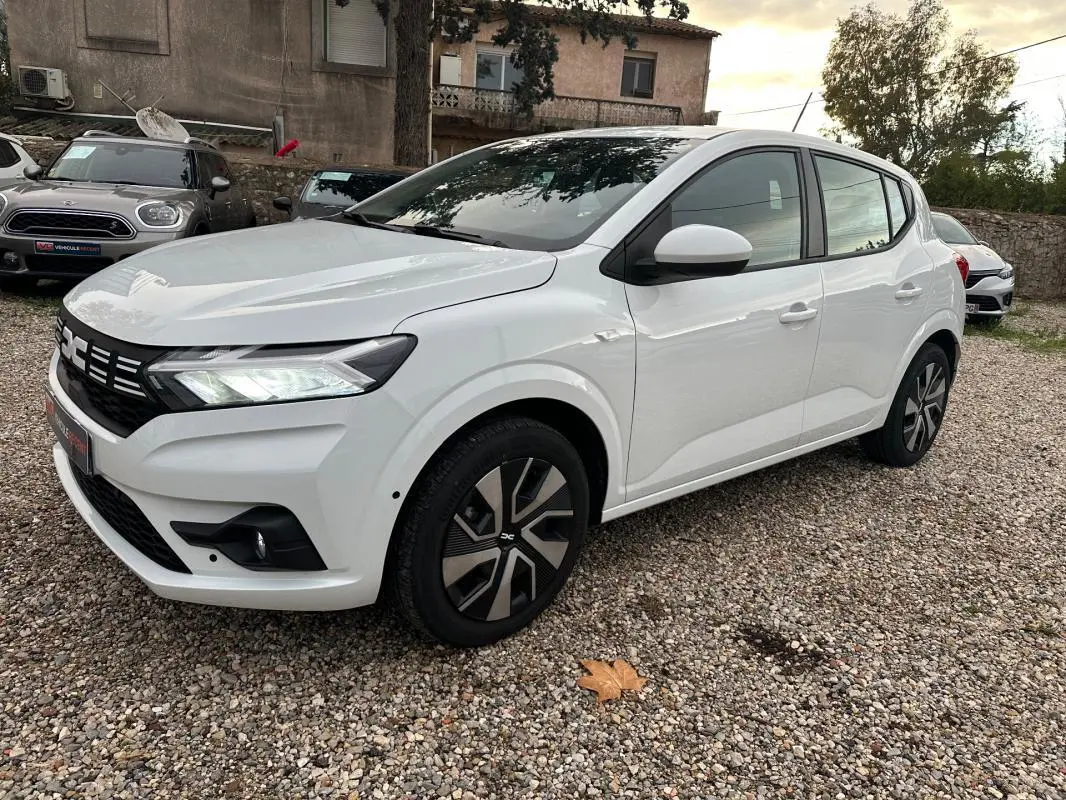 Dacia Sandero TCe 90 blanc vue 3/4 avant droit, avec phares allumés et jantes au design moderne.
