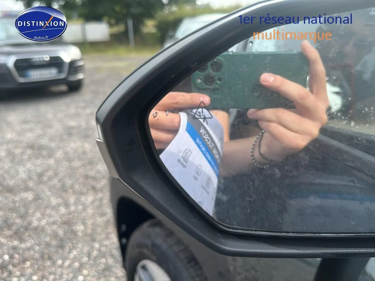 Rétroviseur côté gauche d'un Hyundai Tucson vert cyprès 2025, avec reflet d'une main tenant un smartphone.