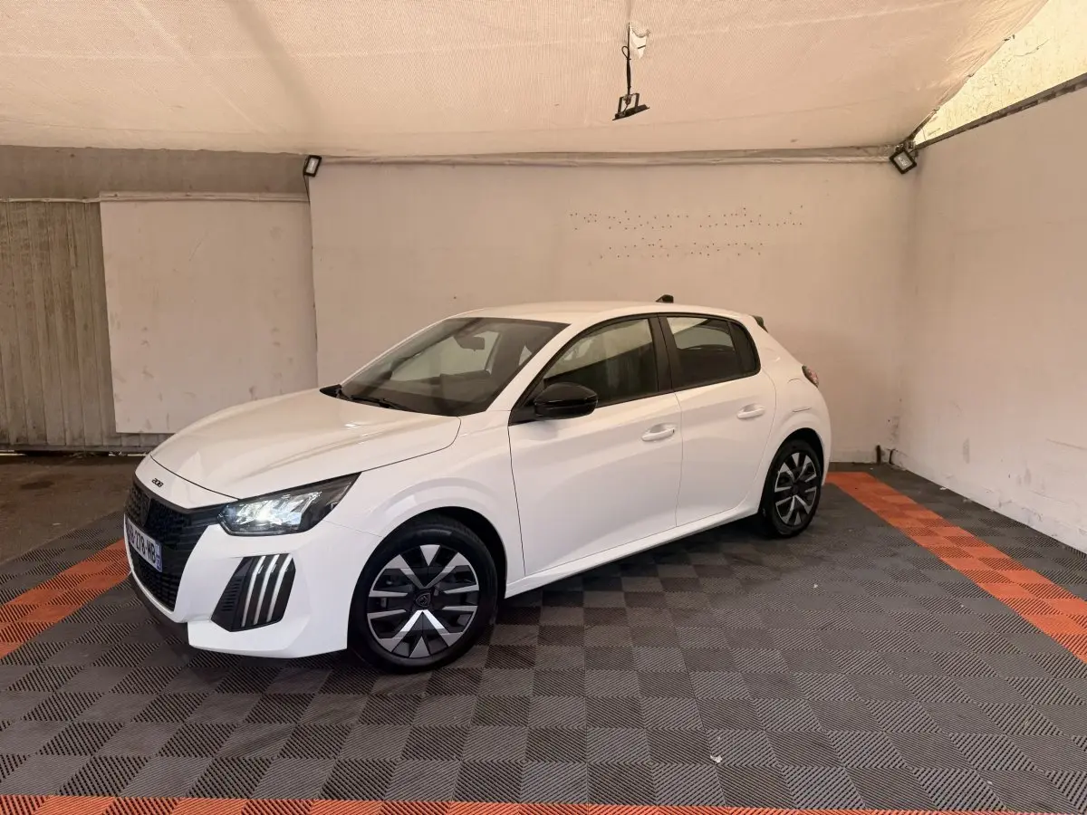Peugeot 208 blanche vue en 3/4 avant droit dans un garage avec jantes noires et phares LED allumés.
