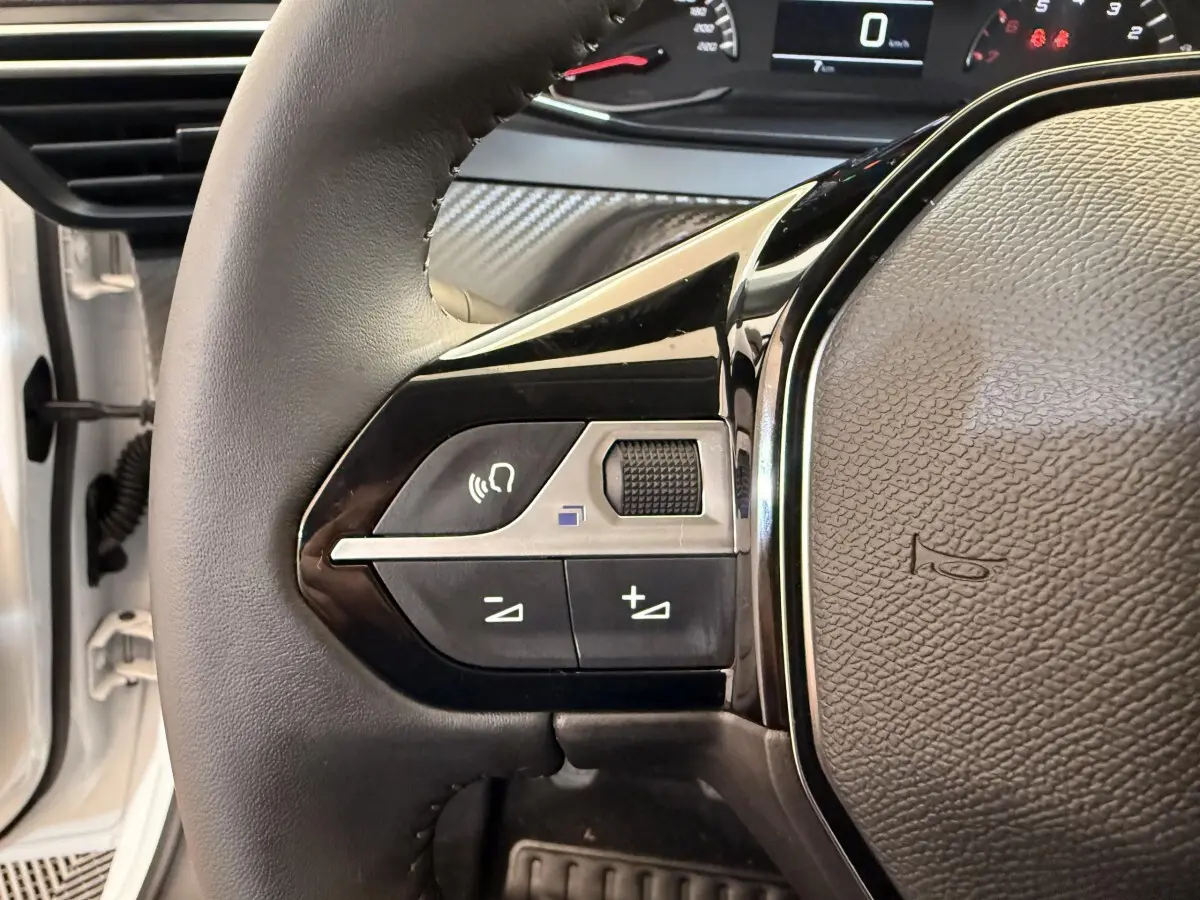 Gros plan sur le volant cuir noir de la Peugeot 208 blanche, avec commandes multifonctions et tableau de bord partiel.