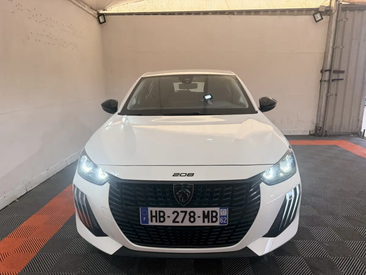 Vue frontale d'une Peugeot 208 blanche 2025 avec phares allumés et calandre noire distinctive.