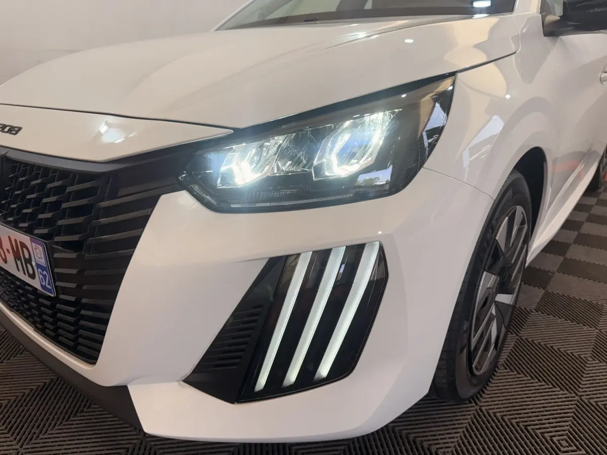 Gros plan sur l'avant droit d'une Peugeot 208 blanche 2025 avec phares LED allumés et signature lumineuse verticale.