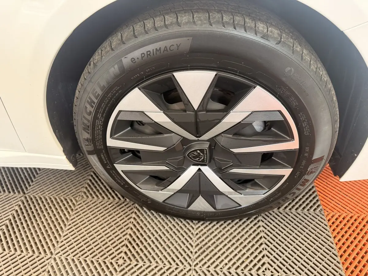 Gros plan sur la roue avant droite blanche d'une Peugeot 208 2025 avec jante au design géométrique noir et argent.
