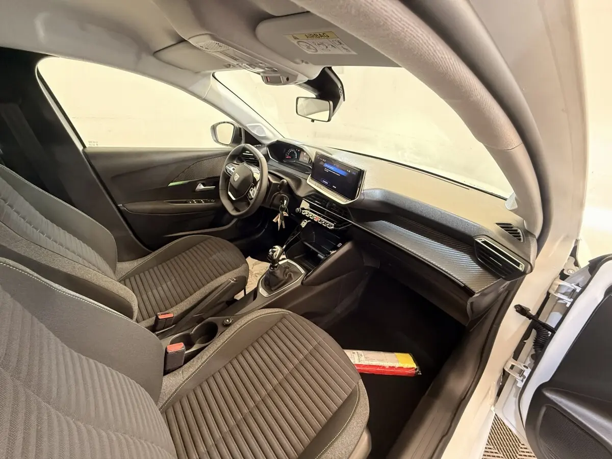 Vue intérieure côté conducteur de la Peugeot 208 blanche 2025, montrant sièges noirs et tableau de bord avec écran tactile.