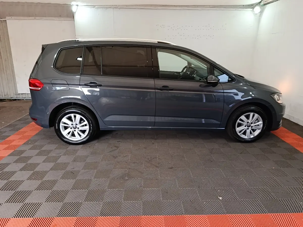 Vue de profil côté gauche d'un Volkswagen Touran gris clair 2024 dans un garage avec sol à damiers noir et orange.