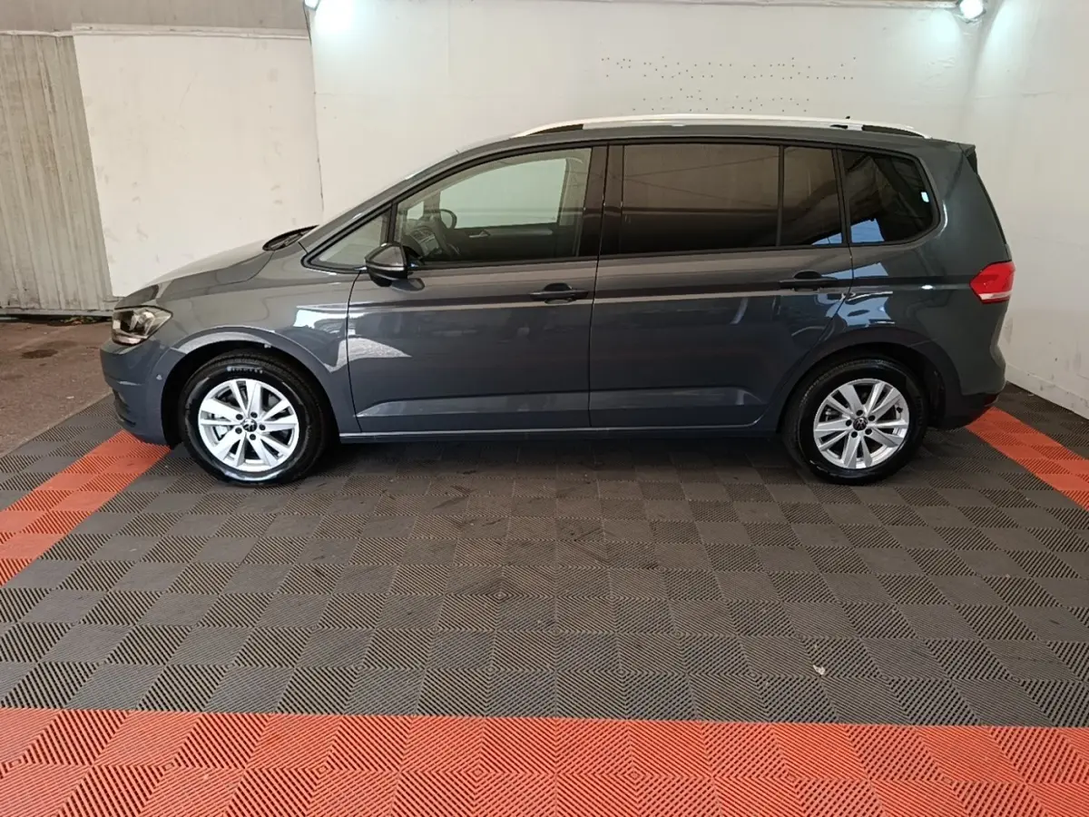 Vue latérale droite d'un Volkswagen Touran gris clair 2024, stationné sur un sol à damiers gris et rouge.