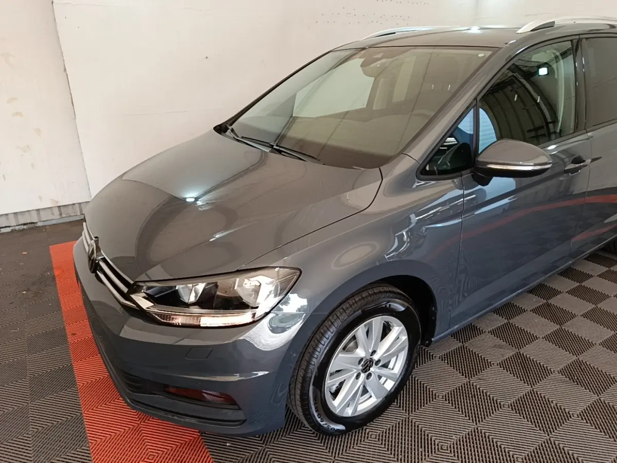 Vue 3/4 avant droit d’un Volkswagen Touran gris clair 2024 avec jantes alliage et feux LED allumés.
