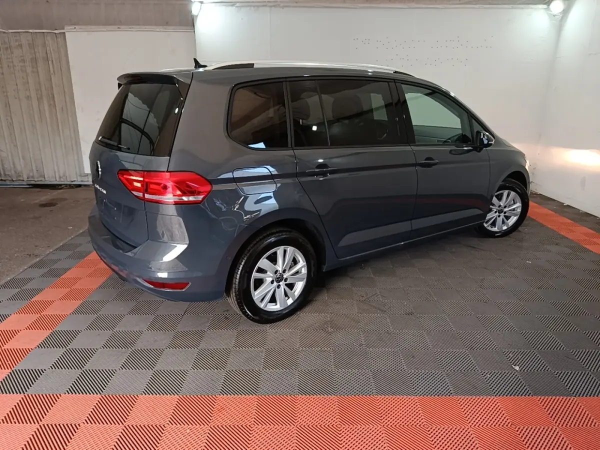 Vue 3/4 arrière droite d'un Volkswagen Touran gris clair 2024 stationné en intérieur sur sol à damiers noir et rouge.