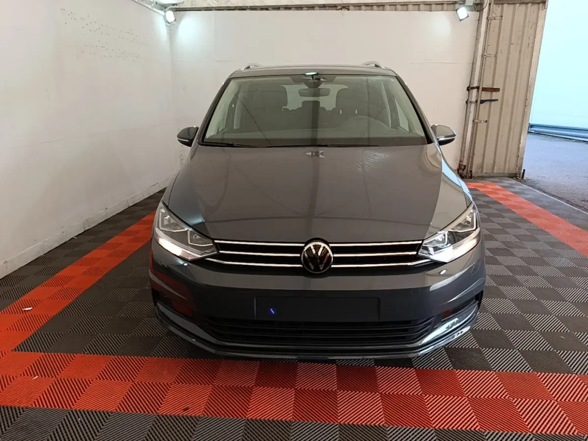 Vue avant d'un Volkswagen Touran gris clair 2024 avec phares allumés dans un garage fermé.