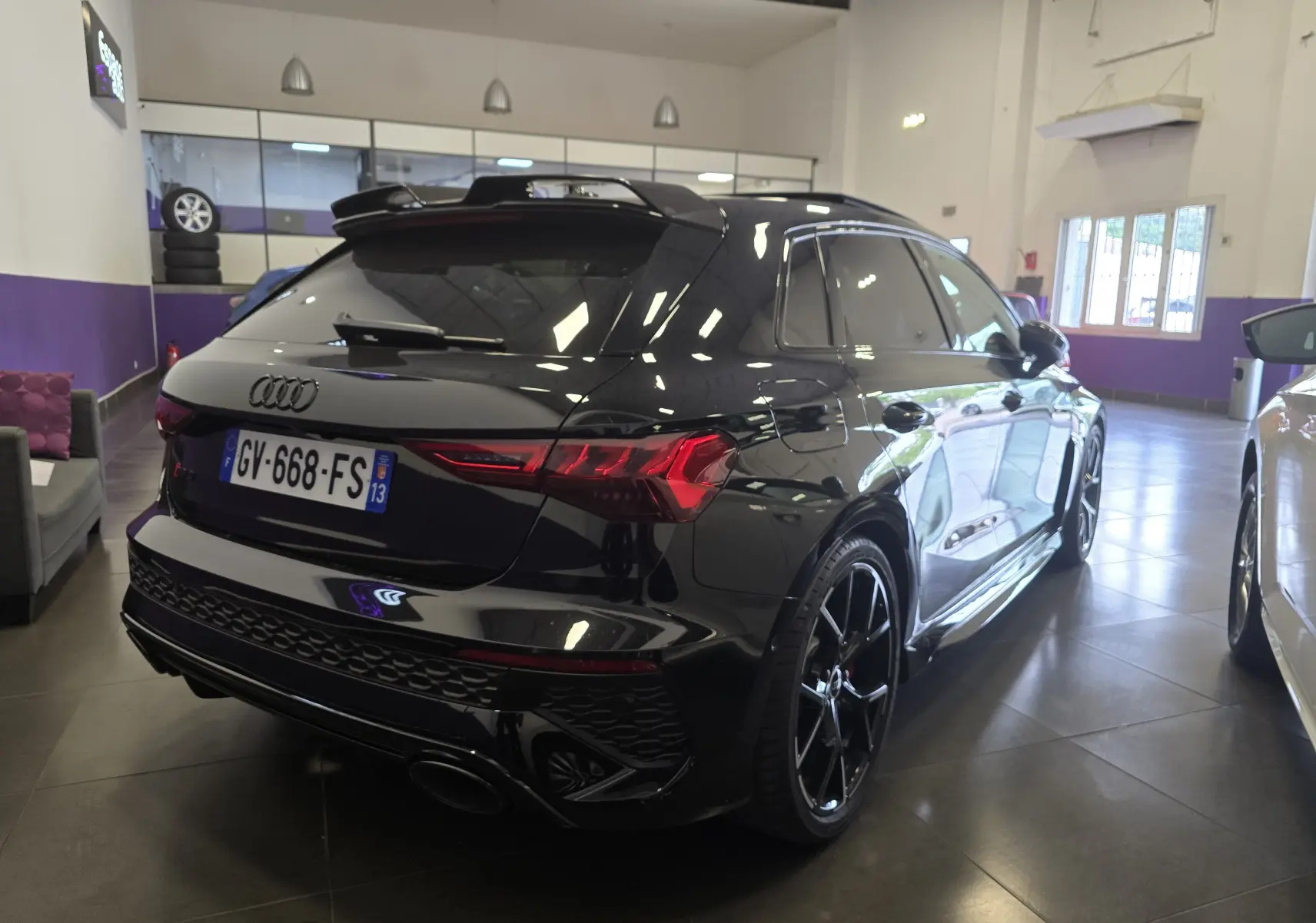Audi RS3 Sportback noir vue 3/4 arrière droit en intérieur, avec jantes noires et feux arrière LED distinctifs.