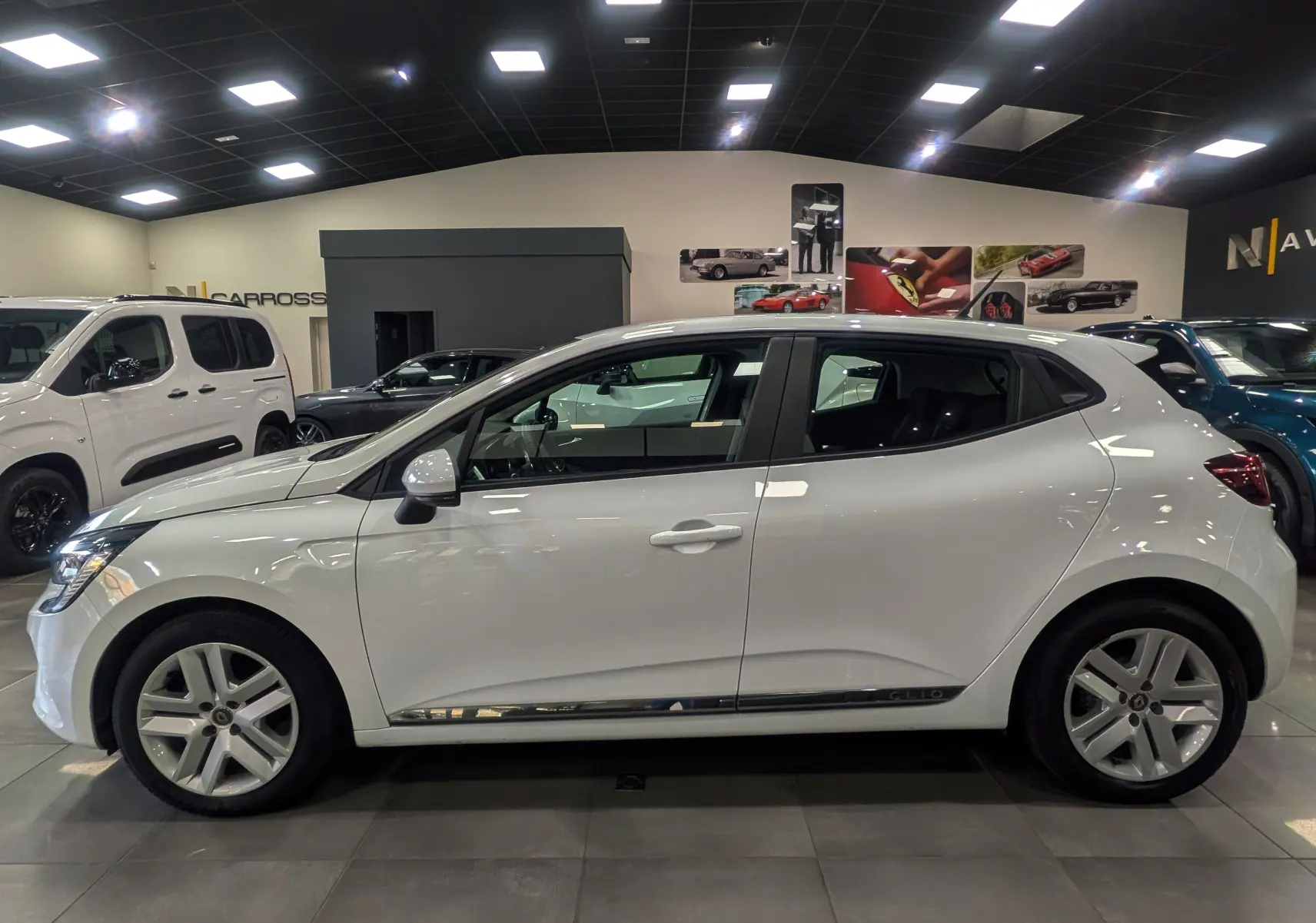 Profil côté gauche d'une Renault Clio V TCe 100 Business blanche Glacier, en intérieur showroom avec jantes alliage.