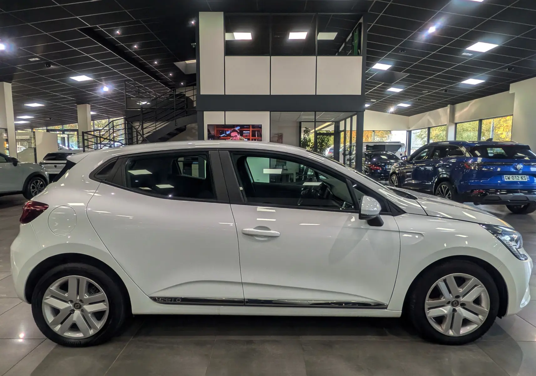 Profil côté gauche d'une Renault Clio V blanche Glacier, version TCe 100 CH BUSINESS, en intérieur showroom.