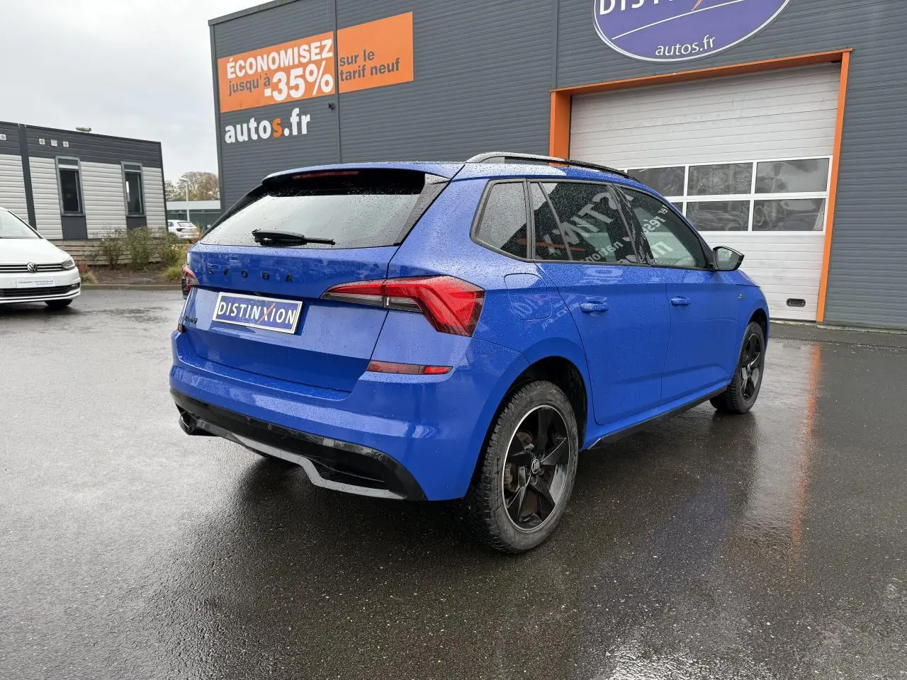 Vue 3/4 arrière droite d'un Skoda Kamiq bleu Energy avec jantes alliage noires et feux arrière LED sur parking humide